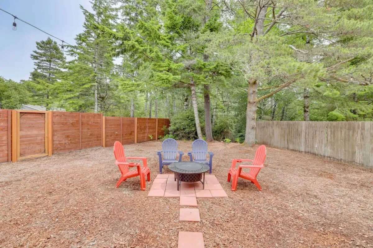 A cozy outdoor fire pit surrounded by red and blue chairs, enclosed by a wooden fence and shaded by mature trees, suggesting a space for gathering, storytelling, or unwinding after a day of coastal adventure.