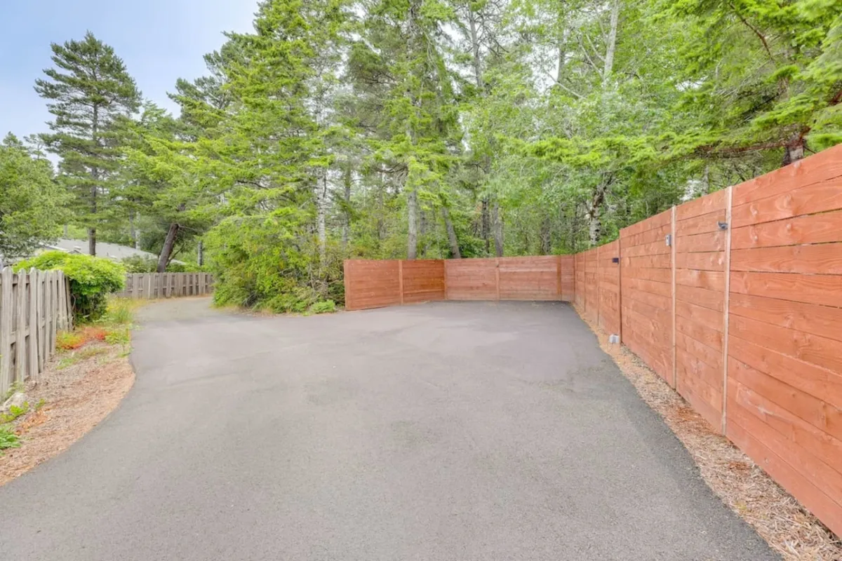 This exterior image shows a paved driveway bordered by a wooden fence and surrounded by lush trees, evoking a sense of natural tranquility. The setting highlights the property's proximity to nature and the great outdoors, hinting at easy access to local trails and the scenic coastal environment described in the section.